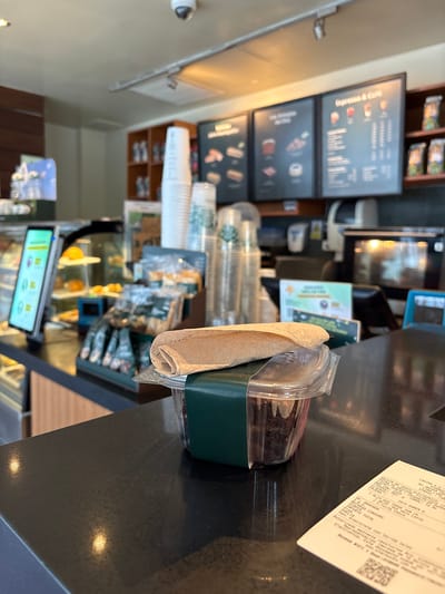 The image shows a close-up of a counter in what appears to be a Starbucks coffee shop in San Isidro, Peru. On the counter, there is a clear plastic container holding a dessert, possibly a chocolate cake or brownie, with a brown paper wrapper placed on top. The container is partially covered by a dark green sleeve. To the right of the container, a receipt is visible, partially laid out on the counter. In the background, there are shelves with merchandise, a menu board displaying food and drink options, and a digital display screen. Stacks of disposable cups are also visible. The overall environment suggests a commercial food and beverage establishment.