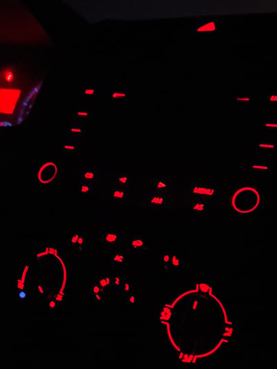 A car's dashboard, illuminated in red, features a variety of controls. On the left are buttons for "MENU" and "AS," along with "AM" and "FM" radio options. Below these are a "CD" button and "TIP". Circular controls for climate settings are visible, including temperature adjustments ranging from 188 to 222, and controls for fan speed (0-4) and airflow direction. Additionally, seat warmer buttons are visible.