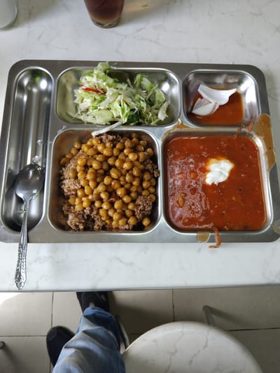 A stainless steel food tray with multiple compartments sits on a marble-patterned surface. In the top left compartment, there is a small portion of what appears to be a salad with shredded lettuce and a few pieces of red pepper. To its right, in a similarly sized compartment, is a small portion of a reddish sauce with two thin slices of white onion. The largest compartment at the bottom is filled with a dish of what looks like rice or buckwheat topped with chickpeas. To the right of that is another compartment containing a reddish stew or soup, garnished with a dollop of white cream or yogurt. A spoon rests in the compartment to the left of the rice and chickpeas. In the background, a glass of dark liquid is visible. The scene is viewed from a slightly elevated perspective, looking down onto the tray and the table. Partially visible in the foreground are a person's legs wearing blue jeans and black shoes, and a white chair. The image is dated to Oliivka, Ukraine.