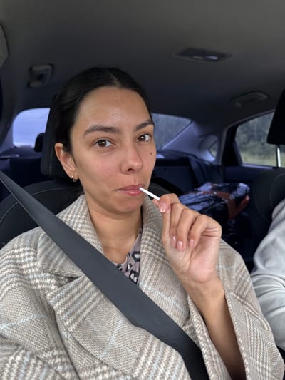 A medium shot shows a woman sitting in the passenger seat of a car, wearing a beige plaid coat over a patterned blouse. She has dark hair, brown eyes, and is holding a white lollipop to her lips. The seatbelt is visible across her chest. The interior of the car is dark, with a glimpse of the outdoor scenery through the window. The location is not specified as Ledukhi, Russia, or any other city.