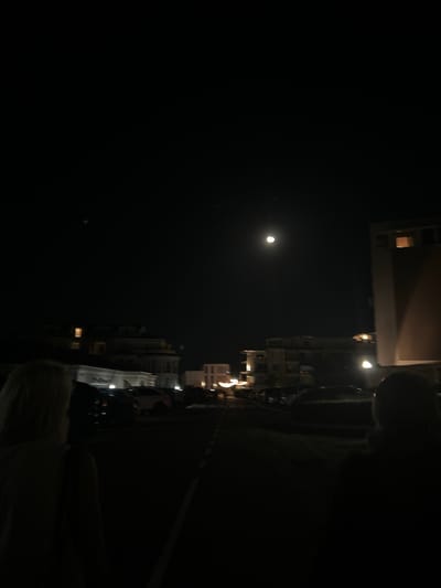 The image shows a night scene in Chernomorets, Bulgaria. The sky is dark with a full moon visible in the upper center. There are several buildings in the background, illuminated by artificial lights. Some of these buildings appear to be apartments or hotels, with lights on in some windows. In the foreground, two figures are partially visible from behind, walking away from the camera. One figure on the left has blonde hair, and the other figure on the right has dark hair. There are cars parked along what appears to be a street or a parking lot. The overall atmosphere is dark and quiet, typical of a nighttime urban or coastal setting.