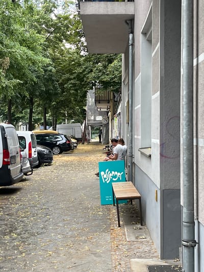 The image depicts a street scene in Berlin, Germany, likely during autumn given the fallen leaves scattered on the ground. In the foreground, a light-colored van is parked, followed by several other vehicles including a silver car, a white compact car, and a black station wagon with a yellow roof rack. Further down the street, a white delivery van is visible, and a person in dark clothing is walking away from the camera. The right side of the image features the exterior of a building with a light-colored facade, marked by vertical architectural elements. Two individuals, both wearing white t-shirts, are seated near a window or opening in the building. A turquoise sign with the word "Passenger" written in white, stylized font is placed on the sidewalk. A wooden bench with dark metal legs is positioned in front of the sign. The ground is paved with a mixture of stone and asphalt, with numerous yellow and brown leaves covering the surface. A metal pipe runs vertically along the side of the building.