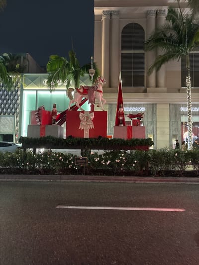 Ferragamo store with holiday decorations. The arrangement includes stacked gift boxes adorned with ribbon, a rocking horse, and a soldier. The boxes are decorated with red and silver ribbon. Lush greenery lines the sidewalk. A lone palm tree is seen in the foreground. The scene appears to be at night, with streetlights illuminating the storefront and decorations.
