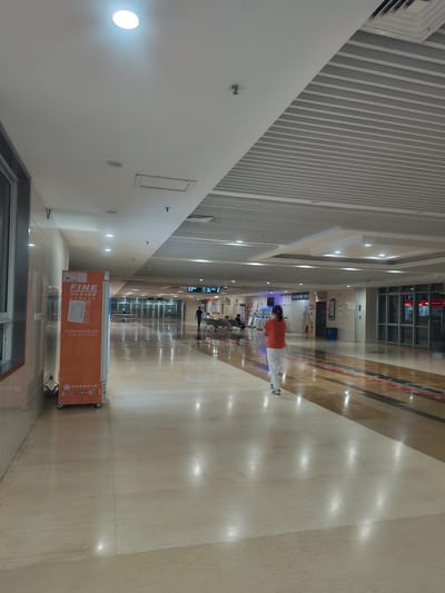 The image depicts a well-lit indoor commercial corridor, likely a shopping mall or airport terminal, in Chongqing High-tech Zone, China. The scene is oriented sideways in the photograph. The corridor features a combination of polished light-colored floor tiles with geometric patterns and a white ceiling with recessed lighting. On the left, a wall is lined with vertical gray and white slats. On the right, the corridor stretches into the distance, with shops or service areas visible through glass facades.

The main subject in the foreground is a person wearing an orange t-shirt and white pants, standing with their back to the camera. They appear to be walking or standing in the middle of the corridor. Further down the corridor, other individuals are visible, some seated and others walking, suggesting a public space with moderate foot traffic.

A prominent orange kiosk or vending machine stands on the right side of the corridor, bearing Chinese text and the brand name "FINE." The visible text on the kiosk translates to "FINE, unmanned vending service, making shopping more convenient" and provides a service hotline.

The overall atmosphere appears to be calm and orderly. The bright, even lighting suggests it is daytime or that the interior lighting is consistently on. There are no obvious indicators of weather conditions. The presence of the vending kiosk and the architectural style of the corridor suggest a modern commercial environment.