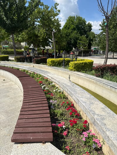 This is a vertical, outdoor photograph featuring a park-like setting with a curved water channel and surrounding flower beds. The water channel, made of concrete with a speckled gray finish, curves from the top right towards the bottom left of the frame. It holds a shallow amount of greenish water. Bordering the water channel, on the side closest to the viewer, is a flower bed with various colorful flowers, primarily red and pink, interspersed with green foliage.
In front of the flower bed is a curved wooden bench, constructed from vertical, dark brown wooden planks. The bench follows the curve of the water channel and flower bed. The ground is paved with light gray concrete slabs.
In the background, several trees with lush green foliage are visible, interspersed with what appear to be buildings. One large tree dominates the left side of the image. There are also some bushes, including bright green, manicured boxwood hedges. Some lampposts or decorative lights are also visible among the bushes.
In the mid-ground and background, several people can be seen. Some are sitting on benches or standing near the bushes and trees. One person appears to be near a bicycle. The activity seems to be leisurely, with people enjoying the park.
The sky in the upper left corner is blue with scattered white clouds, suggesting it is daytime. The lighting indicates a sunny day, likely morning or afternoon. There are no visible text elements in the image. The overall mood is peaceful and pleasant, depicting a well-maintained urban park space.