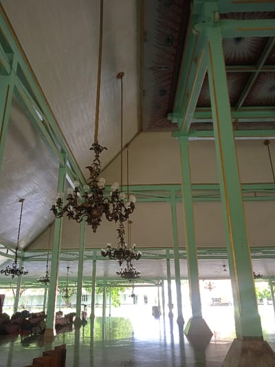 The image shows an interior view of a building. The room has a high ceiling with several large pillars supporting the roof. The ceiling is painted white with green beams. The pillars are painted light green and have gold accents. There are several ornate chandeliers hanging from the ceiling. The room is empty and appears to be a hallway or passageway. The room is well-lit, and the floor is clean and shiny. The image appears to be taken at the Surakarta Palace in Surakarta, Indonesia, located at coordinates -7.559 latitude and 110.783 longitude.