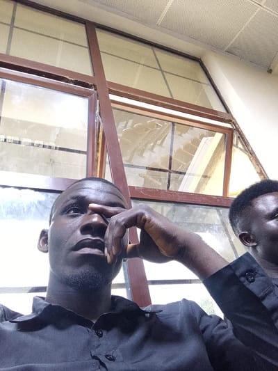 A low-angle, medium shot captures two Black men in the foreground, appearing to be indoors near a window. The man on the left, closest to the camera, is wearing a dark collared shirt. His right hand is raised, with his index finger resting on the bridge of his nose and his thumb near his temple. He is looking upwards and to the left. A faint beard is visible on his chin. To the right and slightly behind him, another man, also wearing a dark collared shirt, is partially visible, facing towards the left and looking down. The background consists of a large window with a reddish-brown frame, divided by vertical and horizontal mullions. The window panes are filled with a textured, translucent material. Beyond the window, the interior of a building is visible, including what appears to be tiered seating. The ceiling above is a white perforated tile grid. The overall lighting suggests an indoor environment, possibly during the day, with the bright window providing some illumination. The location is identified as Kuson, Nigeria.