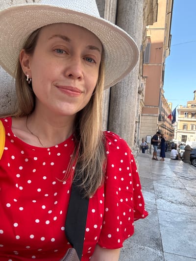The media file shows a woman taking a selfie outdoors, with her face prominently in the foreground. She is wearing a wide-brimmed straw hat and a red dress with white polka dots. She has blonde hair and is looking towards the camera with a slight smile. Her earrings are visible, one of which is a star shape. A thin gold necklace is also visible.
In the background, out of focus, is a street scene in what appears to be a European city. There are people walking, buildings with a classical architectural style, and a cobblestone street. A French flag is visible hanging from one of the buildings, suggesting the location might be an area of French interest or influence. The lighting indicates it is daytime, likely a sunny day given the bright sky. The overall mood seems relaxed and touristy. The image appears to be taken in Rome, Italy, as per the location context.