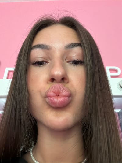 A close-up, waist-up shot shows a young woman with long, dark brown hair against a bright pink background. She is looking directly at the camera and puckering her lips in a kiss. Her eyebrows are well-defined, and she is wearing mascara. She is also wearing a pearl necklace. The background appears to be a wall with some white lettering that is partially obscured. There are no visible elements to indicate the location within Novi Iskar, Bulgaria.