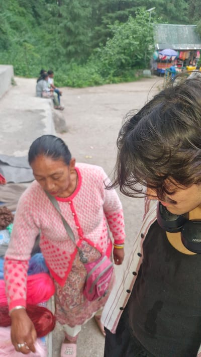 In Manali, India, an elderly woman wearing a pink knitted cardigan over a floral printed dress and white pants is seen attending to a display of colorful yarn. She has a silver ring on her left hand and a yellow bracelet on her right wrist. A small pink crossbody bag is slung over her shoulder. In the foreground, a person with dark, curly hair is partially visible, wearing headphones around their neck and a black t-shirt with a striped jacket over it. The background shows a street scene with lush green trees and a small shop with various items displayed under umbrellas. Two individuals are seated in the distance on a concrete ledge. The ground is a dusty, paved surface.