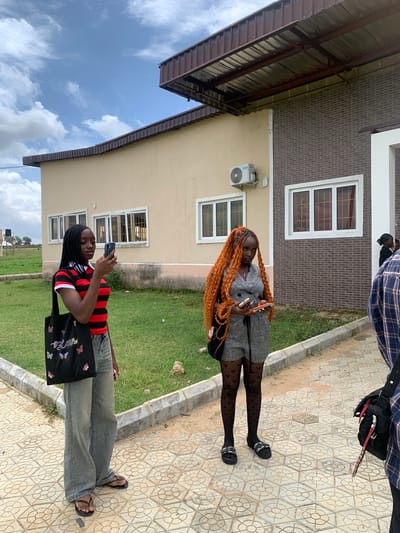 This media file captures an outdoor scene in Gwafan, Nigeria. Two young women are the primary subjects. The first woman, with striking orange braided hair, is standing on a patterned paved walkway, holding two mobile phones. She is wearing a grey patterned dress, black patterned stockings, and black sandals. The second woman is in the foreground, also on the paved walkway, wearing a black and red striped t-shirt, grey wide-leg jeans, and black sandals. She is holding a mobile phone and looking down at it. A black tote bag with butterfly designs and the word "Bliss" is slung over her shoulder.
Behind the women, a modern building with beige and brown brickwork is visible. The building features white-framed windows and an air conditioning unit mounted on the beige wall. A dark metal-roofed overhang extends from the building. To the left of the building, a patch of green grass and a cloudy blue sky are visible.
The image appears to have been taken during the daytime, with natural lighting suggesting it could be late morning or afternoon. The weather seems fair, with scattered clouds in the sky. The paved walkway and the modern architecture suggest an urban or semi-urban environment, possibly a residential or commercial area. The overall mood seems casual and relaxed, with the women engaged with their phones.