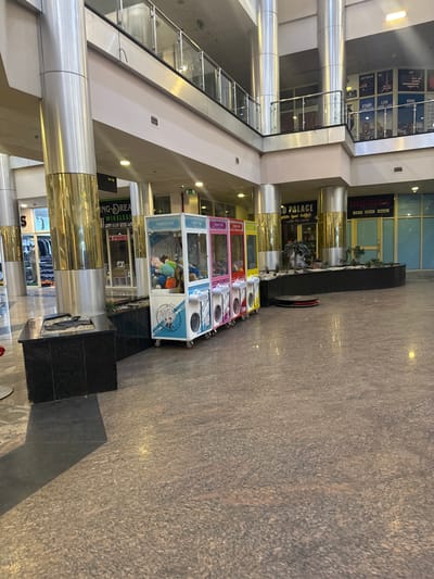 The image depicts the interior of a multi-level commercial building, likely a shopping mall, in Uyo, Nigeria. On the ground level, a row of four "Monster" branded claw crane arcade machines stands near a decorative planter area. The mall features polished stone flooring and metal support columns, with multiple storefronts visible in the background, including one for "ING-DREAM WIRELESS". Upper levels of the mall feature glass railings and additional commercial signage, including a promotional graphic displaying the flags of Australia, the UK, and the USA, along with the words "STUDY ABROAD". The mall appears to be well-lit.