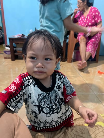 A young child with dark hair and brown eyes is the primary subject in this media file, positioned centrally. They are wearing a colorful shirt with a black collar and intricate black and white patterns, accented with red and white floral designs. To the left and slightly out of focus, an adult, presumably a woman wearing a pink top with white polka dots, is visible. The setting appears to be indoors, with a tiled floor and a light blue wall in the background. There are hints of furniture, including what looks like a wooden chair or shelf. The child's expression is neutral, looking towards the camera. The lighting suggests it is daytime. The crops highlight the child's face, their clothing, and a glimpse of the adult's attire. The overall scene depicts a casual, domestic moment.