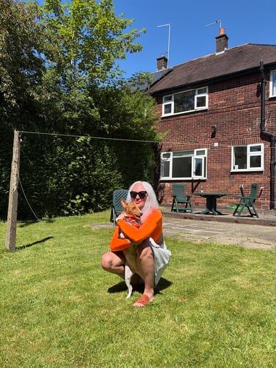 A woman with white hair and sunglasses kneels on a lawn, holding a tan dog wearing an orange garment. The woman is wearing an orange long-sleeved shirt and light-colored shorts. In the background, a brick building with white-framed windows and a dark roof is visible, along with green garden furniture and a dense green tree. The setting is a sunny day, likely in a residential garden in Oldham, United Kingdom. A thin wire extends from a weathered wooden post at the bottom of the frame, suggesting the presence of a clothesline.
