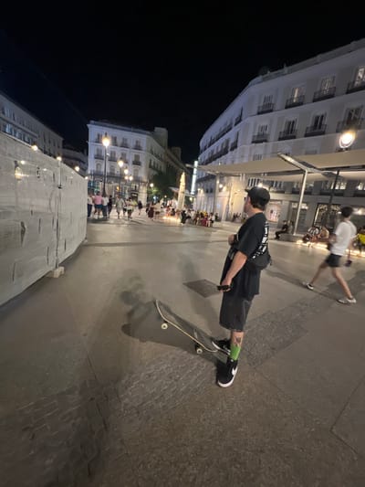 In the image, a young man stands on a paved plaza at night, holding a smartphone in his right hand and a skateboard on the ground to his left. He is wearing a black t-shirt with a graphic design on the back, dark shorts, black sneakers, and bright green socks. A small black bag is slung across his chest. His left leg, adorned with a tattoo, is visible.

The plaza is illuminated by streetlights, casting long shadows. In the background, several people are walking and sitting, suggesting a lively urban environment. To the left of the frame, a large, light-colored barrier, possibly under construction, is covered with a white tarp.

The architecture in the background features multi-story buildings with arched windows and balconies, characteristic of historical European city centers. The presence of awnings over some of the buildings indicates commercial establishments.

The scene appears to be in Madrid, Spain. Specifically, the architecture and the general atmosphere are consistent with the Puerta del Sol, a well-known public square in Madrid. The combination of historical buildings, a bustling plaza, and the presence of street performers or vendors (implied by the gathering of people) are hallmarks of this location.