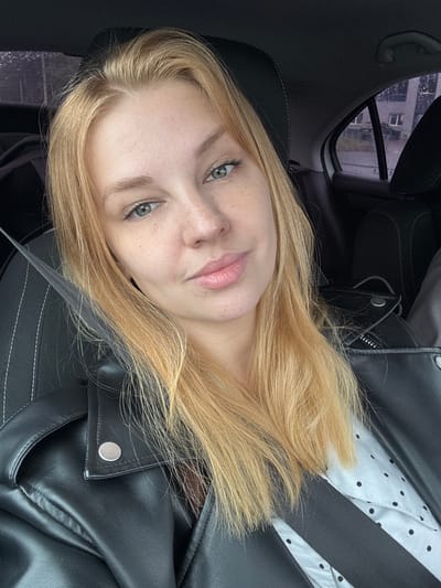 A young woman with blonde hair and light green eyes is sitting in the passenger seat of a car, wearing a black leather jacket over a white polka-dot shirt. The seatbelt is across her chest. It appears to be raining outside, with raindrops visible on the car window, through which a building can be seen. The interior of the car is dark, with black leather seats and headrests. The image is a close-up selfie, focusing on the woman's face and upper body.