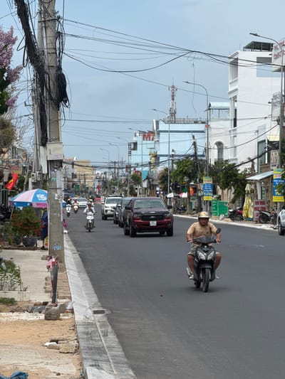 The image depicts a street scene in Phan Thiết, Vietnam, characterized by a relatively narrow asphalt road lined with buildings and dense overhead utility lines. In the foreground, a man wearing a light brown t-shirt and shorts rides a black motorcycle, heading towards the viewer. His face is partially obscured by a helmet. The road is moderately busy with several other motorcycles and cars. To the left of the road, a sidewalk area is visible with a utility pole heavily laden with black wires, a pink and blue umbrella, and some potted plants. Construction debris and sand are also present on the left side. The buildings on the right are multi-story, with a mix of modern white structures and older facades. A cell tower can be seen in the distance. The sky is overcast. The overall impression is one of a bustling, somewhat chaotic urban environment typical of a Vietnamese city.