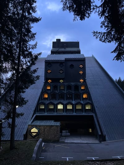 The image depicts the Hotel Bjelasica in Kolašin, Montenegro, under an overcast, deep blue-grey sky. The multi-story building features a prominent, steeply sloped roof or upper facade constructed from dark, possibly metal or slate-like panels. The central part of the building presents a complex, tiered facade with various window configurations. An upper section includes dark-colored panels with hexagonal windows, some of which emit warm yellow light. Below this, multiple rows of hooded, arch-like window alcoves form a textured surface; interior lights, both warm yellow and cool white, are visible in select windows. The lower portion of the facade contains a large, dark recessed area, indicative of an entrance or covered drive. To the left of this recess, a smaller, attached structure with a diamond-patterned exterior features a single illuminated hexagonal window emitting cool white light. The foreground consists of a dark asphalt driveway or parking area marked with white 'T' shapes and an 'X', bordered by a concrete curb and sparse, dark green grass. Dark, silhouetted evergreen trees frame the building on the left and partially obscure the upper right. No persons or active subjects are visible. Some indistinct white objects are discernible within the covered entrance recess.