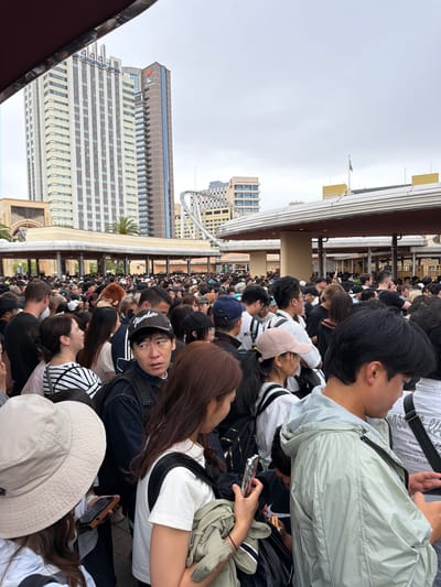A large crowd of people is depicted queuing outdoors, likely at an amusement park or popular attraction. The image shows numerous individuals from various age groups, mostly facing away from the camera, forming a long line that stretches into the distance. Many are dressed casually in t-shirts, jackets, and caps, with some carrying backpacks or shoulder bags. Several individuals are seen holding smartphones, suggesting they are checking messages, taking photos, or passing the time.

In the background, distinctive architectural elements confirm the location as Universal Studios Japan or its immediate vicinity. A prominent skyscraper bears the vertical text "UNIVERSAL STUDIOS JAPAN" along its side, while another building displays the logo for "HOTEL KINTETSU UNIVERSAL CITY." Part of a roller coaster track is visible against the overcast sky, further reinforcing the amusement park setting. An American flag flies from a flagpole in the upper left of the background. The sky is uniformly gray and overcast, indicating a cloudy daytime, possibly cool weather.

Notable details include a man in the foreground wearing a black baseball cap with "Number" written on it. The overall atmosphere appears orderly, with people patiently waiting in line. The crowd density suggests a busy day at the location.