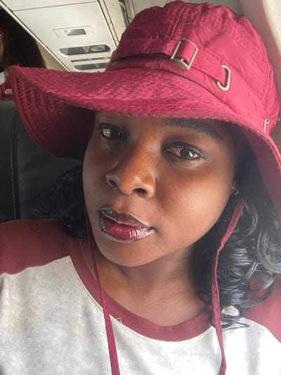 A close-up portrait features a Black woman captured from an angled perspective. She has dark skin and dark, curly hair visible around her face. She wears a maroon or dark red bucket hat with metallic buckles and adjusters on its side, and a matching strap hangs loosely around her neck. Her lips are covered in a glossy, dark red lipstick. She is dressed in a t-shirt that has a white body and maroon trim around the collar, coordinating with her hat. Her gaze is directed confidently towards the camera.
The background is an indoor setting, showing blurred details of what appears to be a vehicle interior, possibly public transport in Ikeja, Nigeria. White panels or cabinets with handles and reflections are discernible, along with a dark surface, potentially a seat. The lighting is bright and even, highlighting the subject's features. No text is visible in the image.