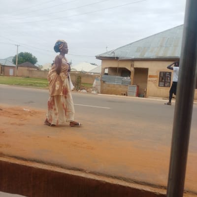 In Gombe, Nigeria, a woman in traditional attire, a patterned headwrap and a light-colored fabric draped over her shoulders and torso, walks along the side of a paved road. Her lower garment is a flowing, cream-colored fabric with large floral patterns in red and pink. She wears dark sandals. The road is bordered by a sandy, reddish-brown shoulder. In the background, a single-story building with a corrugated metal roof is visible, along with other structures and vegetation. To the right, a young man wearing a white t-shirt, black trousers, and sandals, is walking away from the camera, with his right hand touching his head near headphones. Power lines stretch across the cloudy sky. The scene appears to be an outdoor, urban or semi-urban environment.