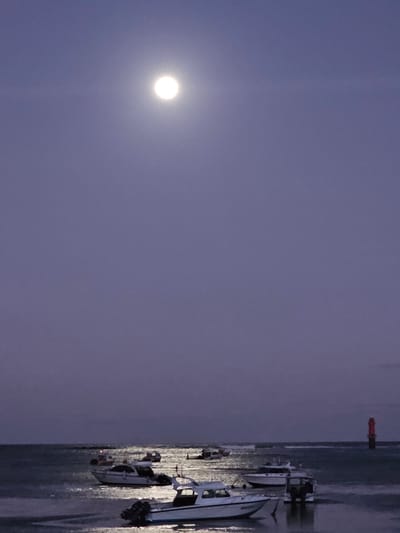 The image depicts a serene night scene on the coast of Denpasar, Indonesia. The sky is a deep purple hue, illuminated by a large, bright full moon situated in the upper left quadrant of the frame. Below the moon, a calm sea stretches to the horizon. Several boats are anchored in the shallow water, their white hulls reflecting the moonlight. In the distance, a red lighthouse stands tall, a solitary beacon against the twilight. The overall impression is one of tranquility and natural beauty.