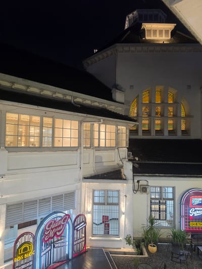 This is a nighttime, eye-level, medium shot of a building in Medan, Indonesia, featuring a mix of architectural styles and illuminated signage. The building itself appears to be old, with some sections showing detailed stained-glass windows and others displaying modern white walls and large pane windows. The overall atmosphere is one of a commercial establishment, possibly a restaurant or cafe, with neon signs contributing to a vibrant, retro feel.
The most prominent objects are the neon signs. One sign, illuminated in red and blue, reads "GUDANG Garasi" and appears to be above a window. Another sign, in blue and pink, says "Garasi" with a stylized font. Below these, a yellow neon sign reads "POS BLOC". These signs suggest a lively and perhaps nostalgic commercial space.
The building's architecture is varied. On the left side, there's a section with a steeply pitched roof and what looks like a large, open window or ventilation shaft. Adjacent to this, a series of arched windows are filled with stained glass in shades of yellow, green, and blue, creating a decorative effect. The right side of the building features large, multi-paned windows that are brightly lit from within, revealing glimpses of interior spaces. There are also potted plants and outdoor seating areas, indicated by dark chairs and tables on a gravel surface, suggesting an outdoor seating area.
The time of day is clearly night, with dark skies and artificial lighting from the signs and interior windows illuminating the scene. The weather appears to be calm. The location cues, such as the text on the signs and the mention of Medan, Indonesia, place the scene specifically. The overall mood is inviting and energetic due to the colorful neon lights. There are no people visible in the image.