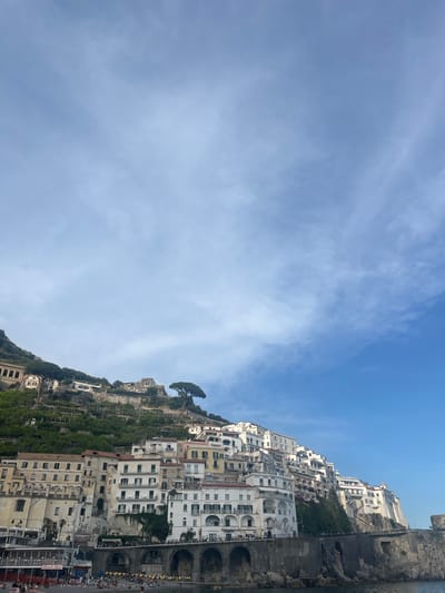 The image depicts the town of Amalfi in the province of Salerno, Campania, Italy, built on a steep hillside. The buildings are primarily white or light-colored, forming dense clusters. Some vegetation, including trees and smaller plants, covers parts of the hillside. The sky above is mostly clear blue with some scattered clouds. The image may be a view from the Pogerola part of the city. The harbor or the bay can be seen on the right with boats and flags.
