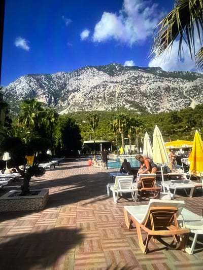 The image depicts a sun-drenched outdoor scene with a focus on a swimming pool area. A large, imposing mountain with a rocky texture dominates the left side of the frame, juxtaposed against a vibrant blue sky with scattered white clouds. Lush green foliage, including palm trees, lines the base of the mountain and extends towards the pool.
The pool area itself is paved with a patterned wooden deck. Numerous lounge chairs, some occupied by people, are arranged around the pool. Several yellow and white umbrellas provide shade. A woman in a dark swimsuit is visible tending to something on a lounge chair. Other individuals are seen relaxing on chairs or walking in the background. The scene suggests a leisurely daytime activity, likely a vacation or a relaxing day at a resort.
The time of day appears to be late afternoon, indicated by the long shadows cast across the wooden deck. The weather is clear and sunny, suitable for outdoor relaxation. While the image does not provide explicit text, the overall atmosphere is one of tranquility and summer enjoyment. The setting appears to be a resort or a hotel with a pool and a view of a mountainous landscape. The presence of lounge chairs, umbrellas, and a swimming pool strongly suggests a leisure-oriented environment.