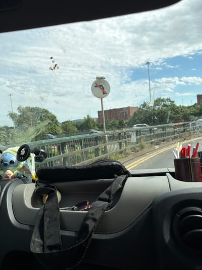 The image is a point-of-view shot from inside a vehicle looking out through the windshield. The windshield is dirty with smudges and what appear to be dried bird droppings. In the foreground, on the dashboard, is a black fabric item, possibly a bag, with a black strap hanging down. To the left, a stuffed toy resembling Stitch from Disney's Lilo & Stitch is visible, attached to a suction cup. To the right, a brown holder contains pens, pencils, and what look like disposable cutlery.
The mid-ground shows a road with yellow lane markings, seen through a green metal railing. Beyond the railing are trees and brick buildings. A traffic sign is visible in the distance, indicating "No Right Turn" with a red circle and diagonal line over a black arrow pointing right. A tall lamppost is also present. The sky is blue with scattered white clouds.
Based on the visual cues, specifically the traffic sign design and the architectural style of the brick buildings, the scene is likely from the United Kingdom. While the image does not contain definitive landmarks of Bristol, the presence of such traffic signage and urban/suburban landscape is consistent with many areas within the city.