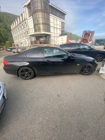 In Listvyanka, Russia, on a paved surface, a black BMW coupe is parked. The car's front passenger side is closest to the viewer. Behind the BMW, a black SUV is parked. In the background, a multi-story building with a sign reading "ОТЕЛЬ ВЕРА" (Hotel Vera) is visible. The sky is blue with some clouds. A wooded hillside is to the left of the building. A portion of a light-colored car is visible on the far left of the image, partially obscuring the BMW.