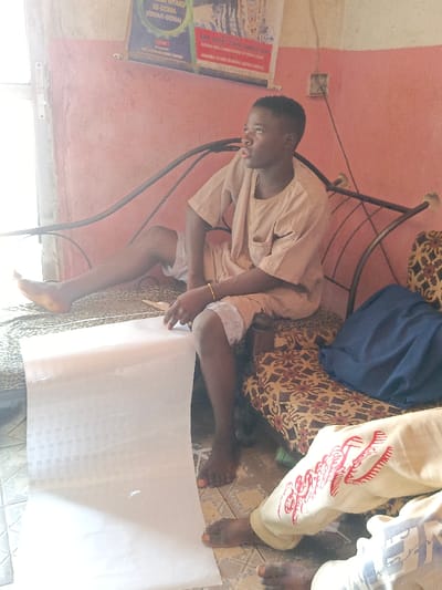 A young man sits on a metal-framed sofa, wearing a short-sleeved shirt and shorts. He holds a roll of white paper, looking to the side with a thoughtful expression. Another person, partially visible, sits beside him, wearing pants with red embroidery. The sofa has patterned cushions and a blue cloth draped over it. The background includes a faded pink wall, a partially visible doorway, and framed posters. The scene is indoors.