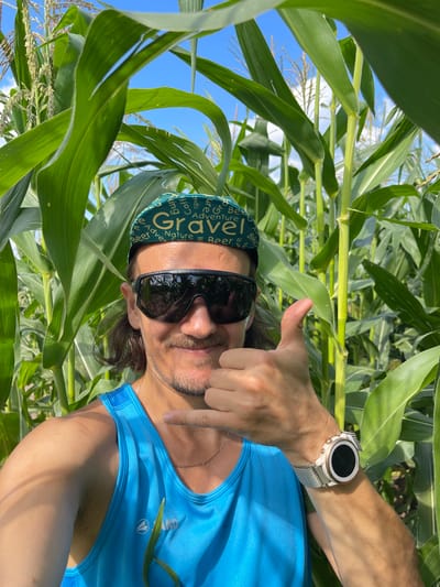 A man wearing a blue tank top, sunglasses, and a green "Gravel" baseball cap is standing in a cornfield. He is making a "shaka" hand gesture with his right hand and gives a thumbs up with his left. The corn stalks are tall and green, and the sky is blue with some clouds. The man has a mustache and stubble. He is also wearing a gold chain and a white smartwatch on his left wrist. The scene appears to be outdoors during the daytime.