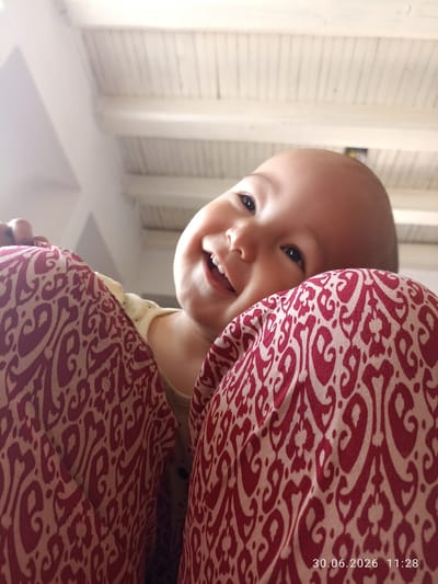 A low-angle close-up shot captures a baby peeking over the shoulder of an adult in Batken City, Kyrgyzstan. The baby, positioned in the upper center of the frame, is looking upwards and smiling broadly, with its mouth open and teeth visible. The adult's shoulder, adorned with a red and white patterned fabric, occupies the foreground, with another portion of the patterned fabric visible on the right side of the frame. The background features a white, slatted ceiling. The date and time "30.06.2026 11:28" are superimposed in the lower right corner of the image.