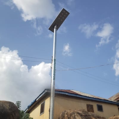 In Gwong, Nigeria, a solar-powered street light stands tall against a blue sky dotted with white clouds. The light, with its rectangular panel of LEDs, is mounted on a silver pole that bears green lettering reading "REAR/DIGITALIZATION/VOT/N150". Below the light, a yellow building with a corrugated metal roof, showing signs of rust and age, is visible. The building has several windows, some boarded up and others with dark panes. In the foreground, large, weathered rocks are scattered, and some greenery can be seen near the base of the building. Power lines are strung across the scene.