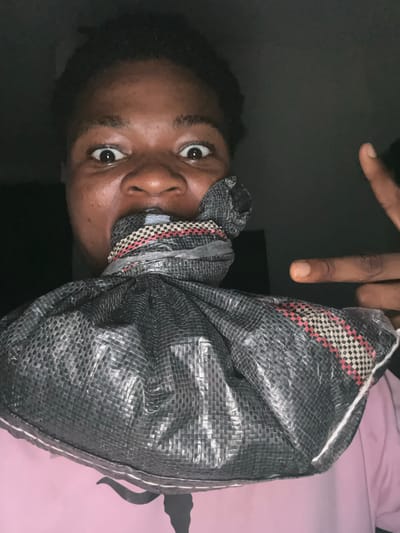 A person with dark skin and dark hair is shown from the chest up, looking directly at the camera with a surprised expression. Their mouth is covered by a black bag that is tied shut. The person is wearing a pink shirt. A finger is raised in the air to the right of their head. The background is dark and indistinct, with no discernible features to indicate a specific location within Ihiagwa, Nigeria.