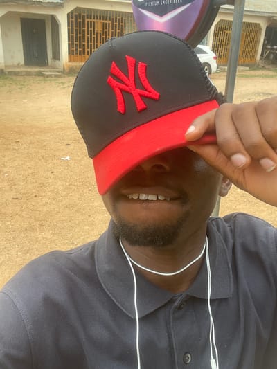 A medium, ground-level outdoor shot depicts a Black man wearing a navy blue button-down shirt and a black baseball cap with a red brim and a red stylized letter on the front. He is holding the brim of the cap with his right hand. White earbud wires are visible around his neck. His face is visible from the nose down, including a beard and a smile revealing white teeth. The background shows a building with a beige exterior and metal window bars. A sign for "Premium Lager Beer" is visible in the top left corner, partially obscuring a white vehicle. The ground appears to be dirt or sand. The location is inferred to be Barakin Akawo, Nigeria.