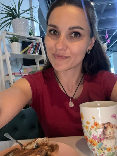 A woman with dark hair and brown eyes, wearing a red t-shirt and a pendant necklace, smiles at the camera. She is seated at a table with a plate of food in front of her and a mug decorated with a rabbit and flowers. In the background, a white shelf holds plants and books. The scene appears to be indoors, possibly a cafe or restaurant.