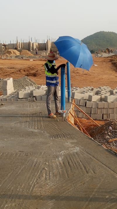 A man in a yellow safety vest and straw hat stands on a concrete slab at a construction site in Zango-ndaji, Nigeria. He holds a blue umbrella and leans against two blue PVC pipes. Stacks of grey concrete blocks surround him. In the background, construction of a low-rise building is underway. Piles of sand and gravel are also visible. A distant, treed hill is in the background. The sky is overcast.