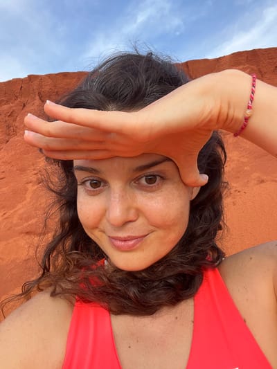 In this close-up selfie, a woman with dark, curly hair is photographed from a low angle, with her head tilted and her face partially obscured by her hand. She wears a vibrant coral-colored top. A red and pink beaded bracelet adorns her wrist. The background features a clear blue sky with scattered clouds and a textured, reddish-orange rock formation, suggesting an outdoor setting. The lighting indicates it is daytime, likely sunny. The woman's expression is a gentle smile, her eyes looking towards the camera. The overall mood is relaxed and cheerful, possibly captured during a vacation or outdoor activity in a warm climate. The location context of Mui Ne, Vietnam, suggests a beach or coastal area known for its sand dunes and scenic beauty, which aligns with the visible landscape elements.