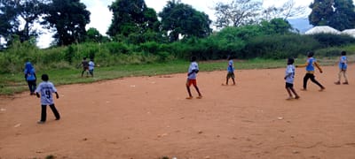 In Ngalula, Democratic Republic of the Congo, a group of children plays on a dirt field. Eight are visible, some wearing soccer jerseys. One jersey has the name "BENZEMA" and the number "9" on the back. In the background, there is a green, dense vegetation and a building with a white roof. Mountains are visible in the distance.