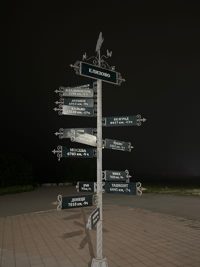 The image shows a signpost with multiple directional signs at night. The signpost is located on a paved area adjacent to a dark road and grassy area. The signpost itself is white and ornately decorated. The signs are dark green with white lettering, indicating various cities and their distances in kilometers and time differences. Some of the visible locations include ЕЛИЗОВО, ВЛАДИВОСТОК, ЛУГАНСК, КАЛЬЯ, МОСКВА, БЕЛГРАД, ТОКИО, МИНСК, ТАШКЕНТ, КРЫМ, ДОНЕЦК, and МАРИУПОЛЬ. The signpost has decorative metal elements and a weather vane at the top. There are no people visible in the image. The lighting suggests an artificial light source illuminating the signpost from the front.