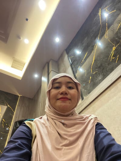 A woman wearing a light pink hijab and a dark blue long-sleeved shirt is taking a selfie. Her face is visible, with makeup on her lips. The background appears to be an indoor setting with marbled walls featuring gold veining and a lit ceiling. The lighting is bright, with several spotlights visible. The image was taken in Banjarbaru, Indonesia.