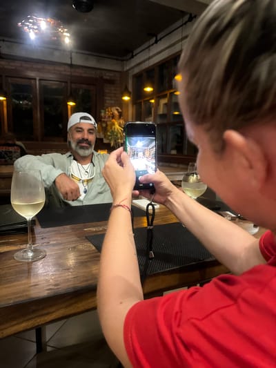 This media file captures an indoor scene at what appears to be a restaurant or bar in Ubud, Indonesia. The primary subjects are two individuals. One person, wearing a red shirt, is in the foreground, holding a smartphone to take a picture. Their arm and shoulder are visible, along with a hint of their hair. The second person is seated at a wooden table, wearing a light-colored jacket and a white baseball cap. They have a beard and are looking towards the person taking the photo, with a slight smile.
The scene is set at a table with two wine glasses, one of which is partially filled with a pale liquid, presumably white wine. The table itself is made of dark, polished wood. In the background, the setting appears to be dimly lit, with exposed brick walls and wooden window frames. Several yellow, glowing lights are visible, adding to the ambiance. One light fixture appears to be a chandelier with multiple bulbs.
The activity taking place is the act of taking a photograph. The person with the phone is framing a shot of the person seated at the table. The overall mood seems relaxed and casual, suggesting a social gathering. There is no visible text in the image. The lighting suggests it is likely evening or nighttime. The style of the interior, particularly the brickwork and wooden elements, might be typical of restaurants in Southeast Asia, aligning with the provided location context of Ubud, Indonesia.