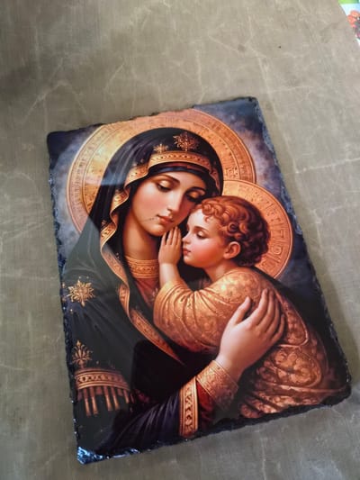 The image depicts a religious icon of the Virgin Mary and the infant Jesus, rendered in a realistic style with rich colors and intricate details. The Virgin Mary is portrayed with a dark blue veil adorned with gold trim and ornamentation, while the infant Jesus is dressed in a golden, patterned garment. Both figures have halos, indicating their divine status. The icon is set against a backdrop that features a circular, gilded motif with what appear to be inscribed characters. The overall composition evokes a sense of tenderness and maternal love. The icon is placed on a textured surface, likely a fabric or mat, which has a subtle pattern of diagonal lines. The edges of the icon appear to be uneven and somewhat rough, suggesting it might be made of stone or a similar material. The lighting in the image is soft, casting gentle shadows and highlighting the textures and details of the icon. The background is a muted, neutral color, which helps the vibrant colors of the icon to stand out. There is no discernible urban or natural environment present in the image. The location of Kalipetrovo, Bulgaria is not depicted or suggested by any visual elements within the provided image.
