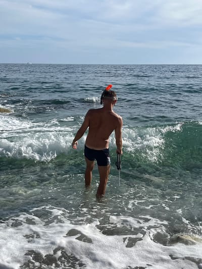 A man stands in the shallow, clear water near a rocky shore, preparing to swim or dive. He is wearing dark swim trunks and a snorkel mask with an orange mouthpiece. He holds a speargun in his right hand, pointing downwards. Waves with white foam are breaking around him. The sea is a deep blue-green, and the sky above is mostly clear with some clouds. In the distance, a small white boat is visible on the horizon. The scene appears to be a natural, undeveloped coastal area, potentially near Bar, Montenegro, given the common activities and scenery associated with the Adriatic coast.