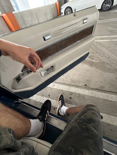 A first-person perspective shot captures a person's hands and legs from a seated position inside a car, with the car door open. The car interior features a cream-colored door panel with a wood grain accent and chrome trim. The person's left hand is visible, reaching towards the car's interior door handle mechanism. The person is wearing camouflage-patterned shorts, white socks, and black Adidas sneakers. The exterior view shows a concrete parking lot with white parking lines and a white car parked nearby. The overall lighting suggests an outdoor, daytime scene, possibly in a parking garage or an open-air lot, which could be located in Los Angeles, California, United States of America.