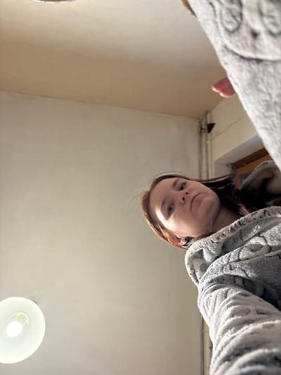 A low-angle shot presents a woman with reddish-brown hair, wearing a gray patterned fleece jacket, looking downward. Part of a light-colored textured surface is visible at the top left. A bright light fixture illuminates a plain, light-colored ceiling. The location is potentially indoors, but there is no specific evidence in the image to confirm it is in Murino, Russia.