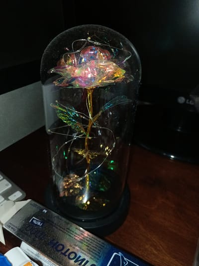 The image displays a decorative artificial rose encased in a glass dome, illuminated by tiny fairy lights. The rose appears to be made of iridescent material, reflecting various colors. The glass dome has a slight film on its surface, suggesting it may be dusty. The dome sits on a black circular base.

In the foreground, to the left, a portion of a product box with Russian text is visible. The text on the box includes "ИМОТОК 1000", "гель", and "M". Further left, a silver keyboard is partially visible. The background is dark, with a hint of a dark surface that might be a desk. A portion of an LG electronic device with its logo is also visible on the right side of the image. The lighting suggests an indoor setting, likely during the evening or in a dimly lit room, as the fairy lights are prominent. There are no people in the image. The overall scene evokes a sense of decorative display or personal adornment.