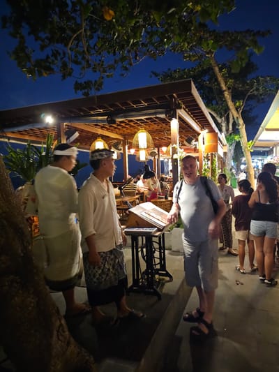 This media file depicts an outdoor scene at night, likely a restaurant or a similar establishment in Denpasar, Indonesia. The primary subjects are several people, some standing and some seated. In the foreground, two individuals are seen from the waist up, wearing traditional Balinese attire with white shirts and headbands, suggesting a cultural event or performance. One of them is looking upwards with a serene expression.

Further back, a man in a grey t-shirt and shorts is standing near a podium with a menu or information displayed on it, and he is looking towards the camera with a slight smile. Several other people, mostly women, are visible in the background, some appearing to be patrons of the establishment. The setting features a rustic, open-air structure with wooden beams and lattice work, illuminated by warm, ambient lighting from hanging lamps with woven shades. Tropical foliage, including trees with large branches, is visible on the left side of the image, adding to the outdoor ambiance. A vintage sewing machine-like object is placed near the podium.

The time of day is clearly evening or night, indicated by the dark blue sky and the artificial lighting. The weather appears to be pleasant, suitable for an outdoor gathering. The overall mood is relaxed and social. There is no visible text in the image that is clearly legible.